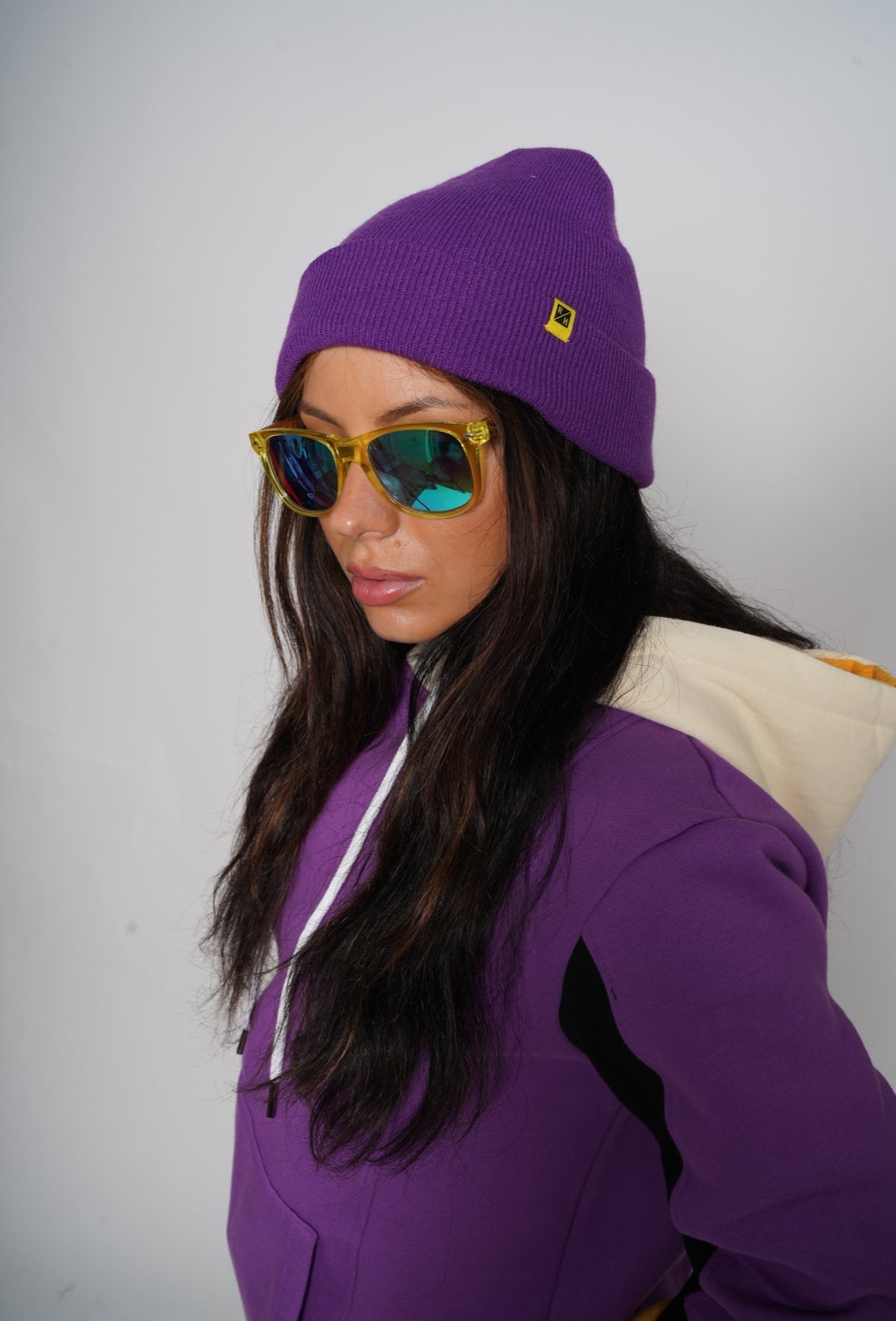 A woman wearing a purple knit beanie with a small yellow logo, yellow sunglasses with mirrored lenses, and a purple hoodie with a cream hood, photographed against a plain light gray background.