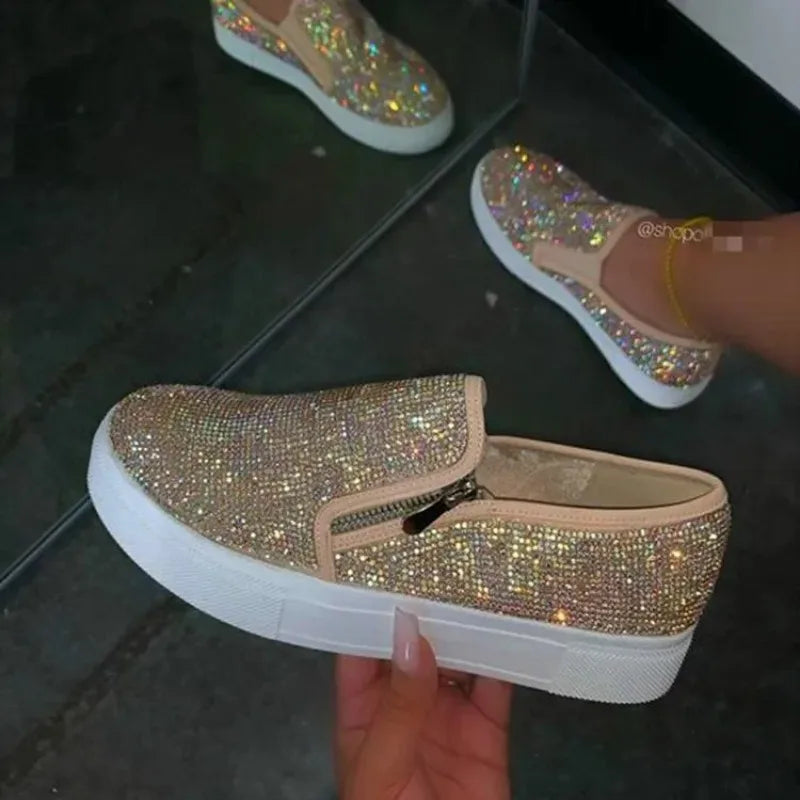 Glittery slip-on shoes with a white sole held by a hand on a reflective surface.