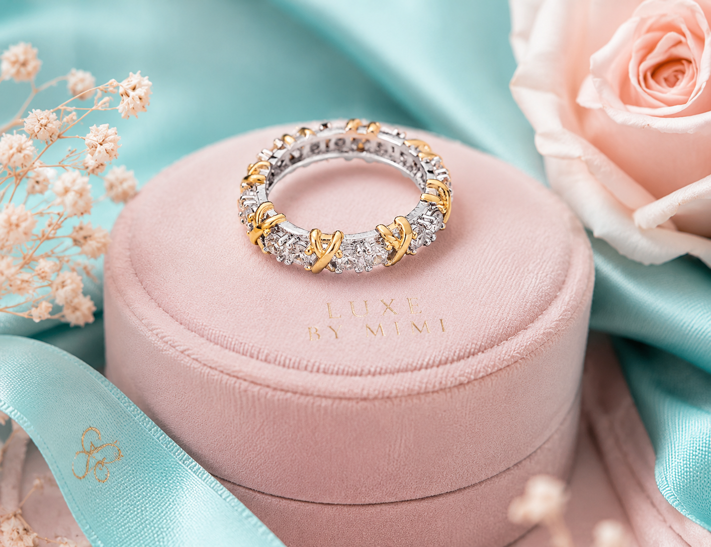 Gold and silver ring on a pink cushion with 'Luxe by Mimi' branding, surrounded by flowers.