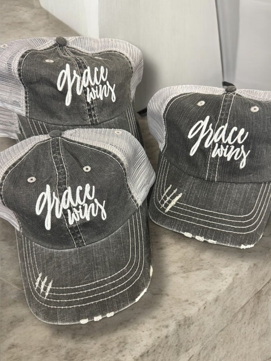 Three distressed baseball caps with 'Grace Wins' text on a marble surface. Grace Wins Embroidered Trucker Hat – Inspirational Christian Faith Adjustable Mesh Cap for Men & Women