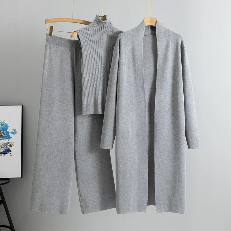 Gray knit outfit with cardigan, sweater, and pants on hangers against a gray wall.