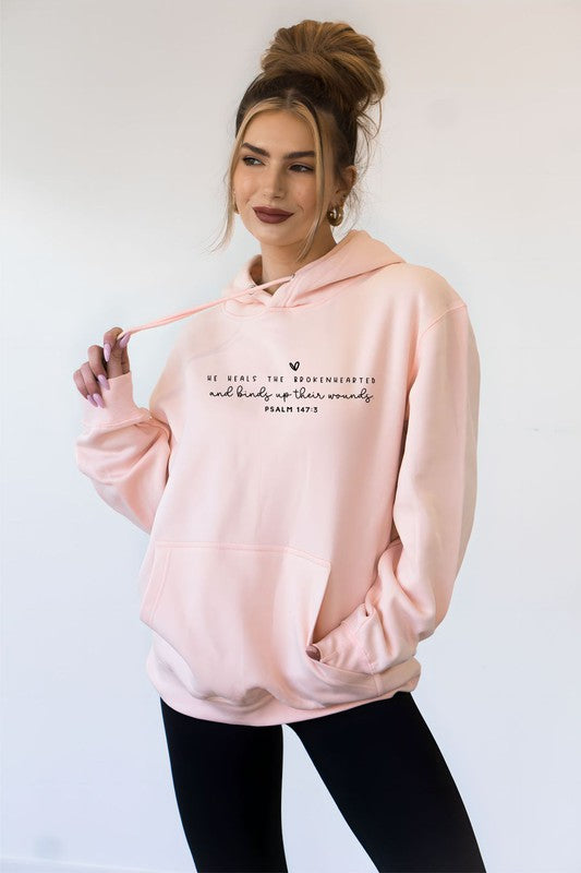A smiling woman wearing a soft pink Christian hoodie featuring the Bible verse "He heals the brokenhearted and binds up their wounds" from Psalm 147:3, posing against a plain white background.