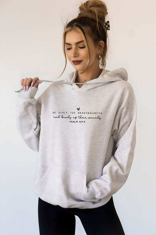 A smiling woman wearing an oatmeal heather Christian hoodie featuring the Bible verse "He Heals the Brokenhearted and Binds Up Their Wounds" (Psalm 147:3), holding the hoodie drawstring while posing against a plain white background.