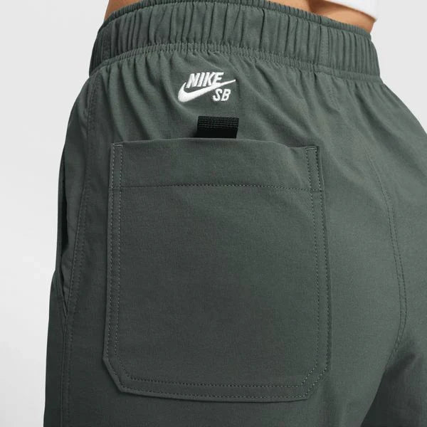 Close-up of dark gray Nike SB shorts showing the elastic waistband with a white embroidered logo, a side pocket, and a black hook-and-loop strap detail. These lightweight, sweat-wicking pants are made for movement and comfort. With 6 pockets