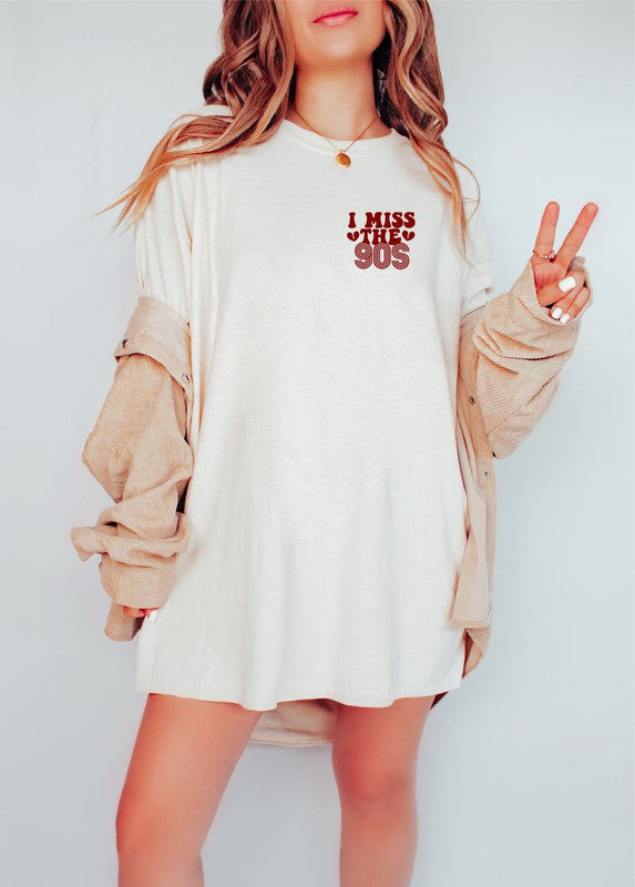 Woman wearing a cream softstyle graphic T-shirt with a vintage red "I Miss The 90s" left chest print, styled with a beige oversized jacket and making a peace sign.