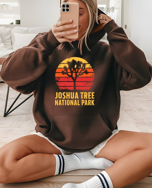 Person wearing a brown hoodie with 'Joshua Tree National Park' design, sitting on a couch. Joshua Tree National Park Graphic Hoodie Material: 80% ring-spun cotton, 20% polyester.