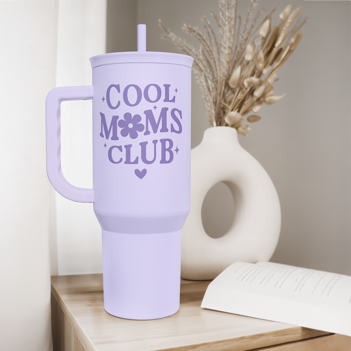 Lavender tumbler with 'Cool Moms Club' text on a wooden surface with a vase and book in the background.