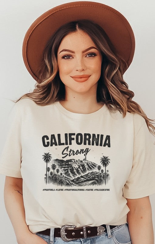 Woman wearing a beige **California Strong Hollywood** graphic unisex T-shirt with a vintage Los Angeles skyline and palm tree design, paired with blue jeans and a brown wide-brim hat against a neutral background.