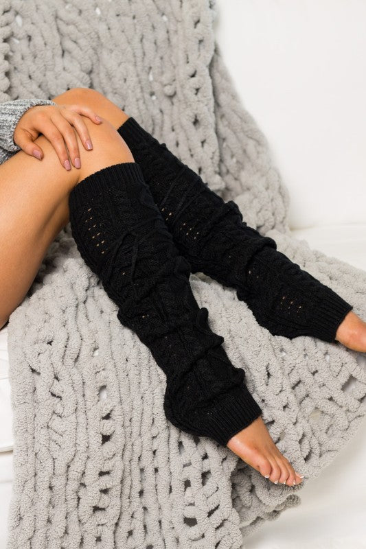 Woman lounging on a white sofa wearing black cable knit leg warmers featuring decorative lace-up tie accents and a textured over-the-knee design. These cozy knitted leg warmers offer stylish cold-weather layering and pair perfectly with boots, leggings, dresses, skirts, and oversized sweaters. Crafted with a thick cable knit pattern for warmth and comfort, the versatile leg warmers can be worn slouched or extended for a chic winter fashion look. Ideal for fall and winter outfits, cabin getaways, lounging at