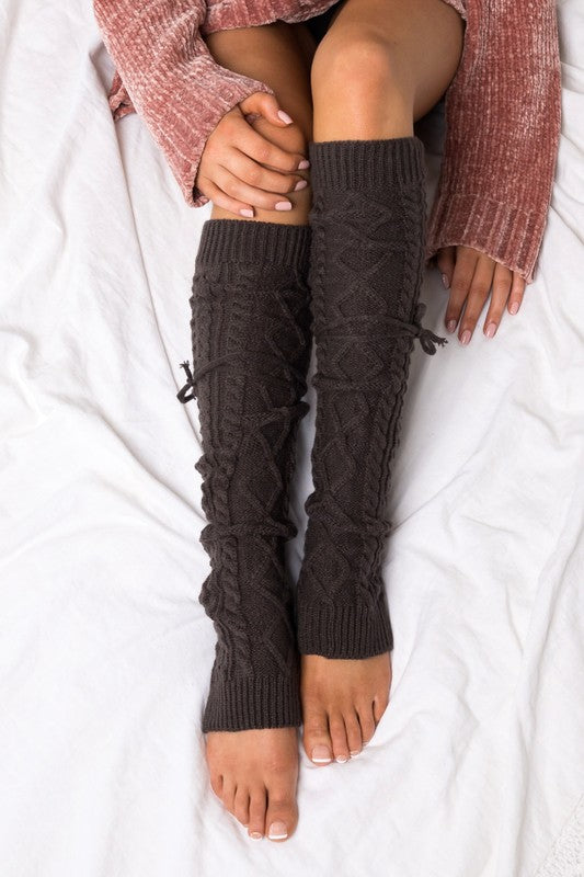 Woman lounging on a white sofa wearing black cable knit leg warmers featuring decorative lace-up tie accents and a textured over-the-knee design. These cozy knitted leg warmers offer stylish cold-weather layering and pair perfectly with boots, leggings, dresses, skirts, and oversized sweaters. Crafted with a thick cable knit pattern for warmth and comfort, the versatile leg warmers can be worn slouched or extended for a chic winter fashion look. Ideal for fall and winter outfits, cabin getaways, lounging at