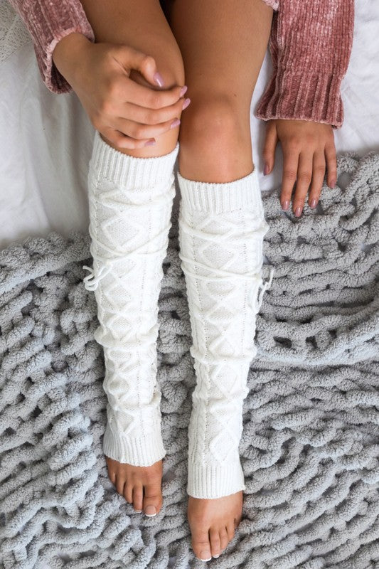 Woman lounging on a white sofa wearing white cable knit leg warmers featuring decorative lace-up tie accents and a textured over-the-knee design. These cozy knitted leg warmers offer stylish cold-weather layering and pair perfectly with boots, leggings, dresses, skirts, and oversized sweaters. Crafted with a thick cable knit pattern for warmth and comfort, the versatile leg warmers can be worn slouched or extended for a chic winter fashion look. Ideal for fall and winter outfits, cabin getaways, lounging at