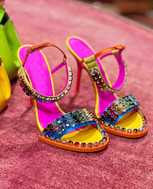 Colorful rhinestone high-heel sandals with rainbow crystal embellishments, vibrant pink insoles, orange soles, ankle straps, and open-toe design displayed on a soft pink velvet surface.