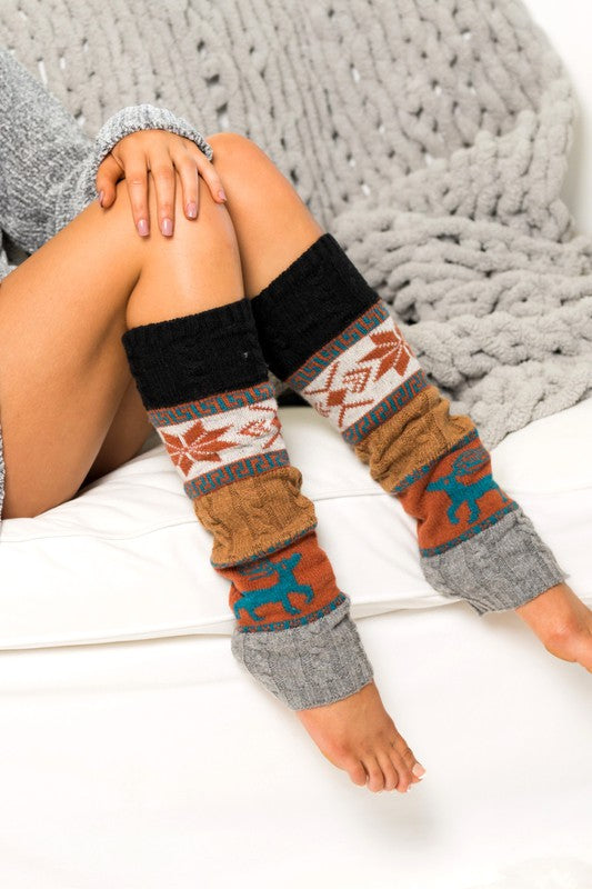 Woman wearing teal Nordic snowflake leg warmers featuring a traditional winter Fair Isle pattern with snowflakes, geometric accents, and deer motifs in gold, gray, and navy tones. These cozy 100% soft-spun acrylic leg warmers showcase a thick knitted construction with color-block detailing and ribbed cuffs for warmth and comfort. Styled while lounging indoors beside a chunky knit blanket, these fashionable leg warmers are perfect for pairing with boots, leggings, jeans, tights, and winter outfits. Ideal for