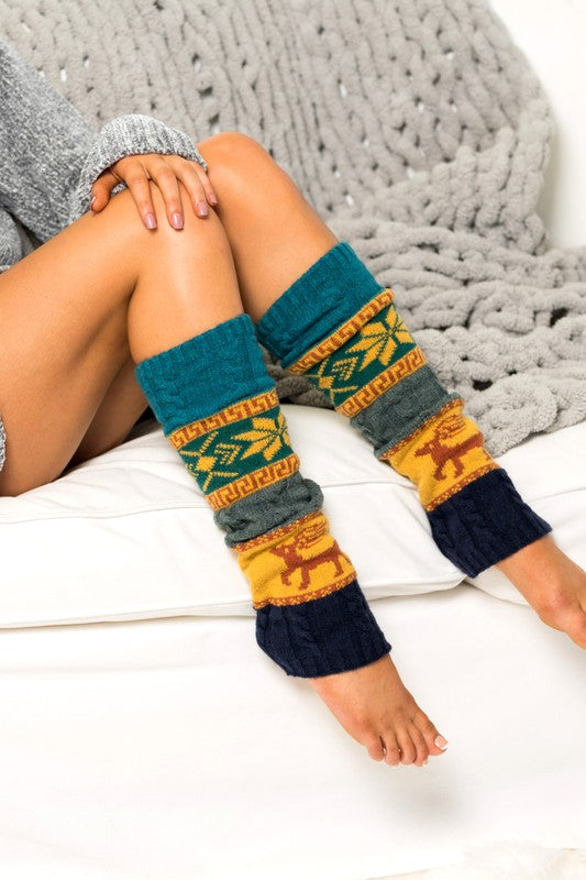 Woman wearing teal Nordic snowflake leg warmers featuring a traditional winter Fair Isle pattern with snowflakes, geometric accents, and deer motifs in gold, gray, and navy tones. These cozy 100% soft-spun acrylic leg warmers showcase a thick knitted construction with color-block detailing and ribbed cuffs for warmth and comfort. Styled while lounging indoors beside a chunky knit blanket, these fashionable leg warmers are perfect for pairing with boots, leggings, jeans, tights, and winter outfits.