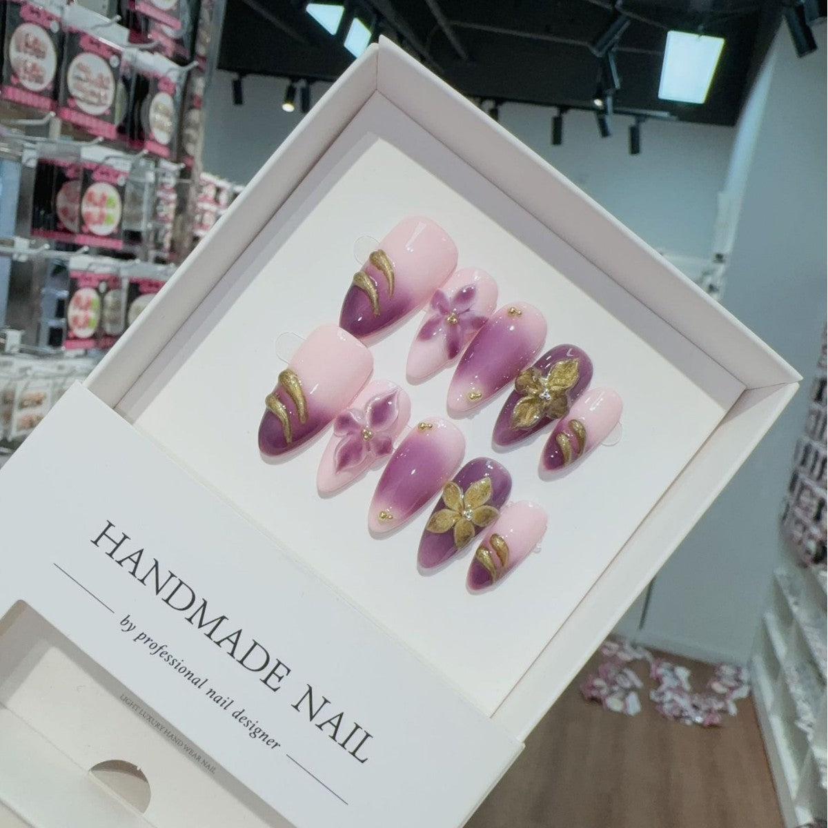Box of handmade press-on nail art with pink and purple designs in a store setting