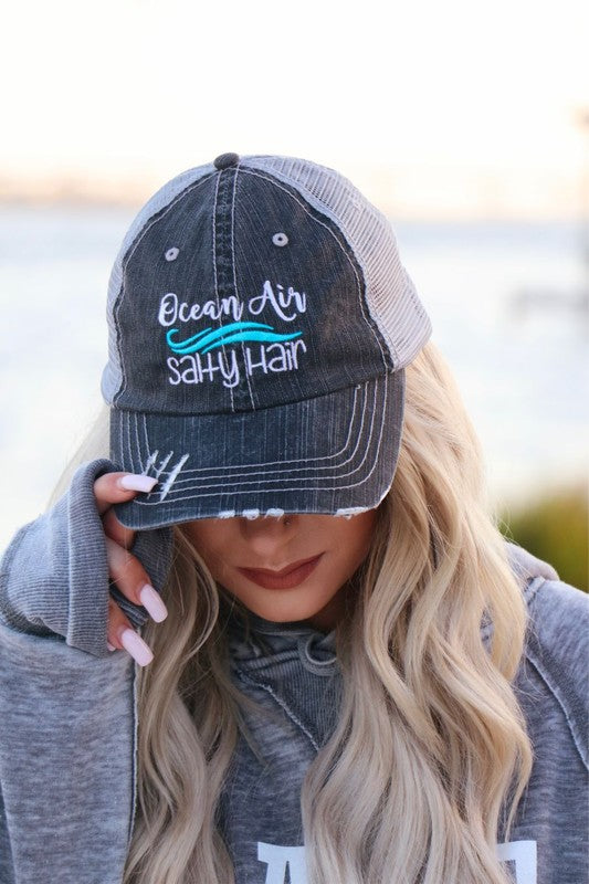 Person wearing a cap with 'Ocean Air Salty Hair' text, standing by a body of water.
