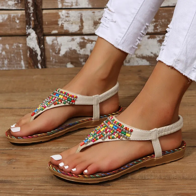 Women's PU leather flat thong sandals with colorful beaded embellishments, elastic slingback strap, braided trim sole, and open-toe design, shown in beige with white distressed jeans on a rustic wooden floor.