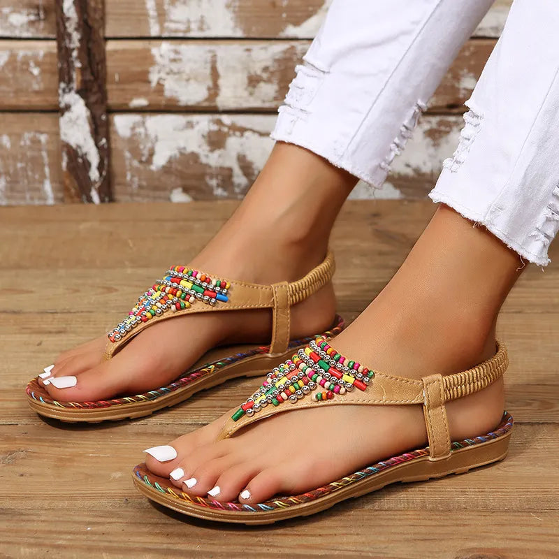 Women's beige PU leather flat thong sandals with colorful beaded embellishments, elastic slingback strap, braided trim sole, and cushioned footbed, worn with white distressed jeans on a rustic wooden floor.