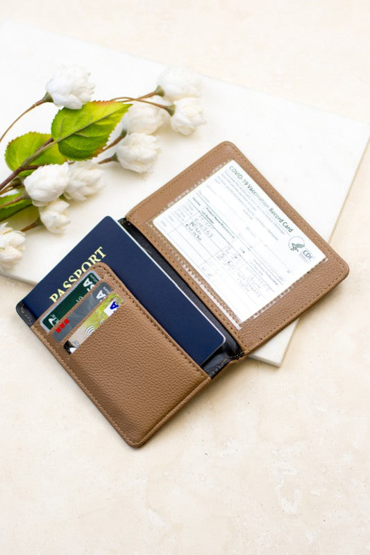 Tan passport and vaccine card holder wallet with passport, credit cards, and vaccination record displayed in multiple slots on a light neutral background with white cotton flowers.