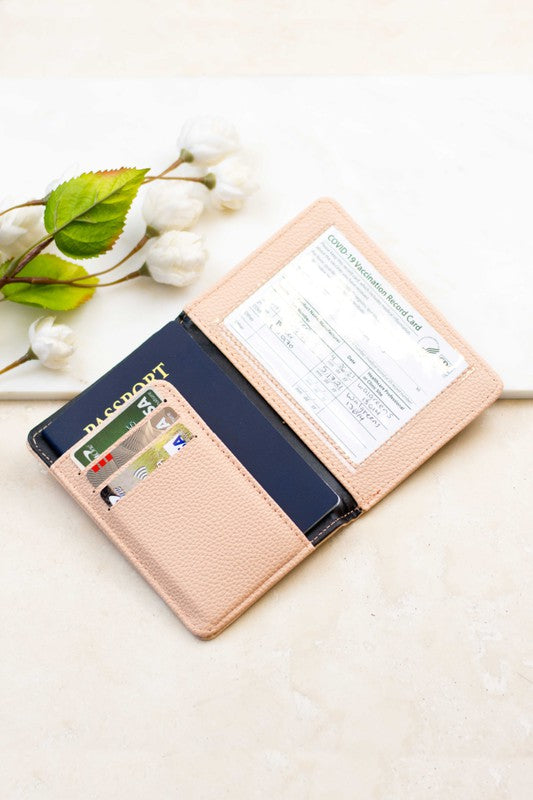 Blush pink passport and vaccine card wallet with card slots, holding a passport, payment cards, and vaccination record on a neutral background with white flowers.