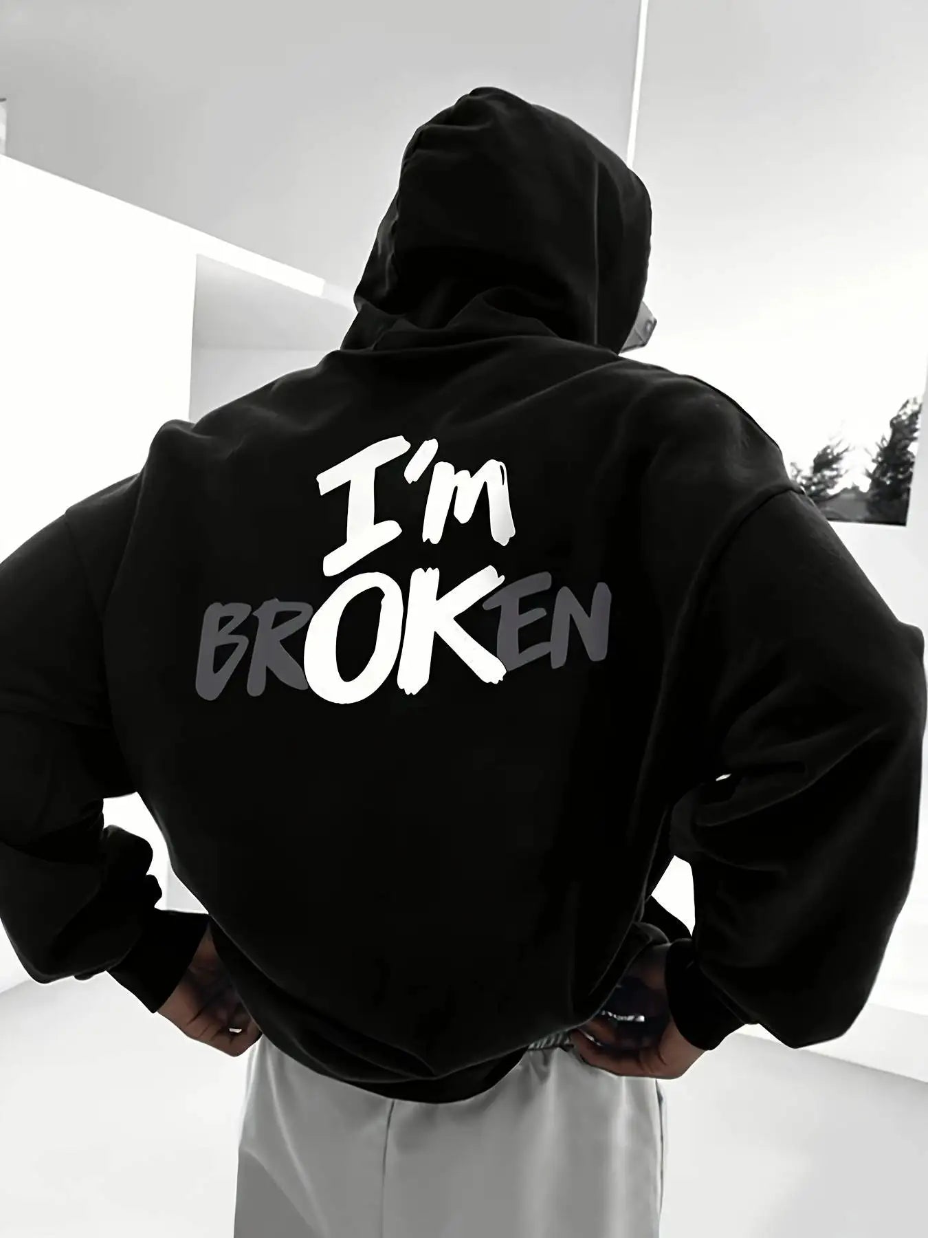 Person wearing a black hoodie with 'I'm Broken' text on the back, standing in a minimalistic setting.