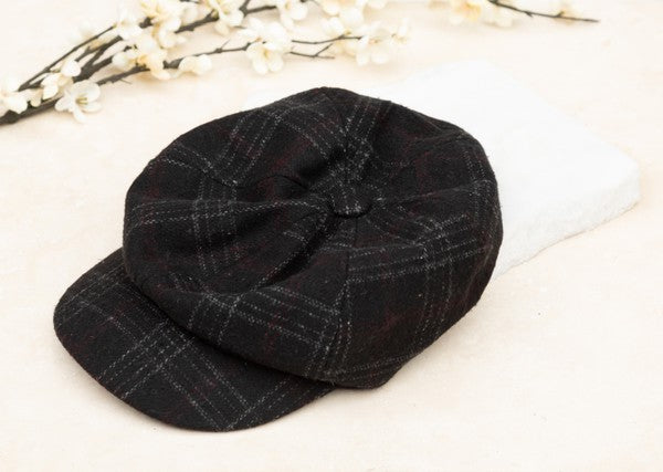 Black plaid newsboy cap with a vintage-inspired checkered pattern, soft structured crown, gathered button top, and short curved brim displayed on a light fabric background.