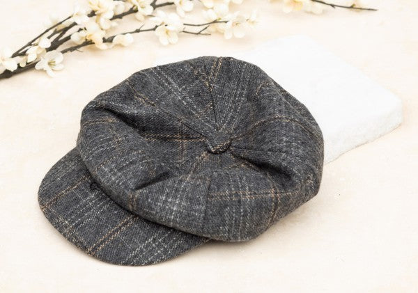Brown plaid newsboy cap with a classic vintage-inspired design, featuring a structured crown, button top, and short curved brim, displayed on a light fabric background.