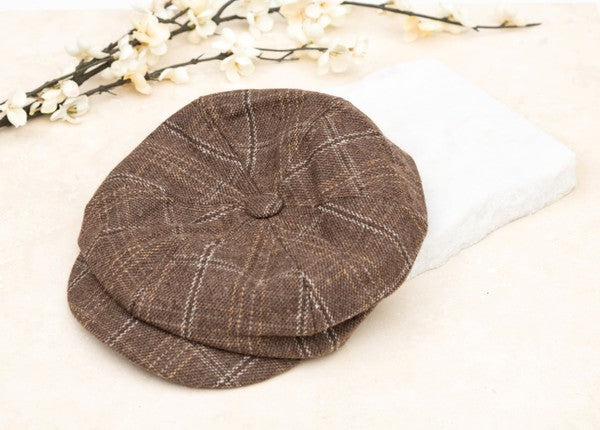 Brown plaid newsboy cap with a vintage-inspired checkered pattern, structured crown, button top, and short curved brim displayed on a light fabric background.