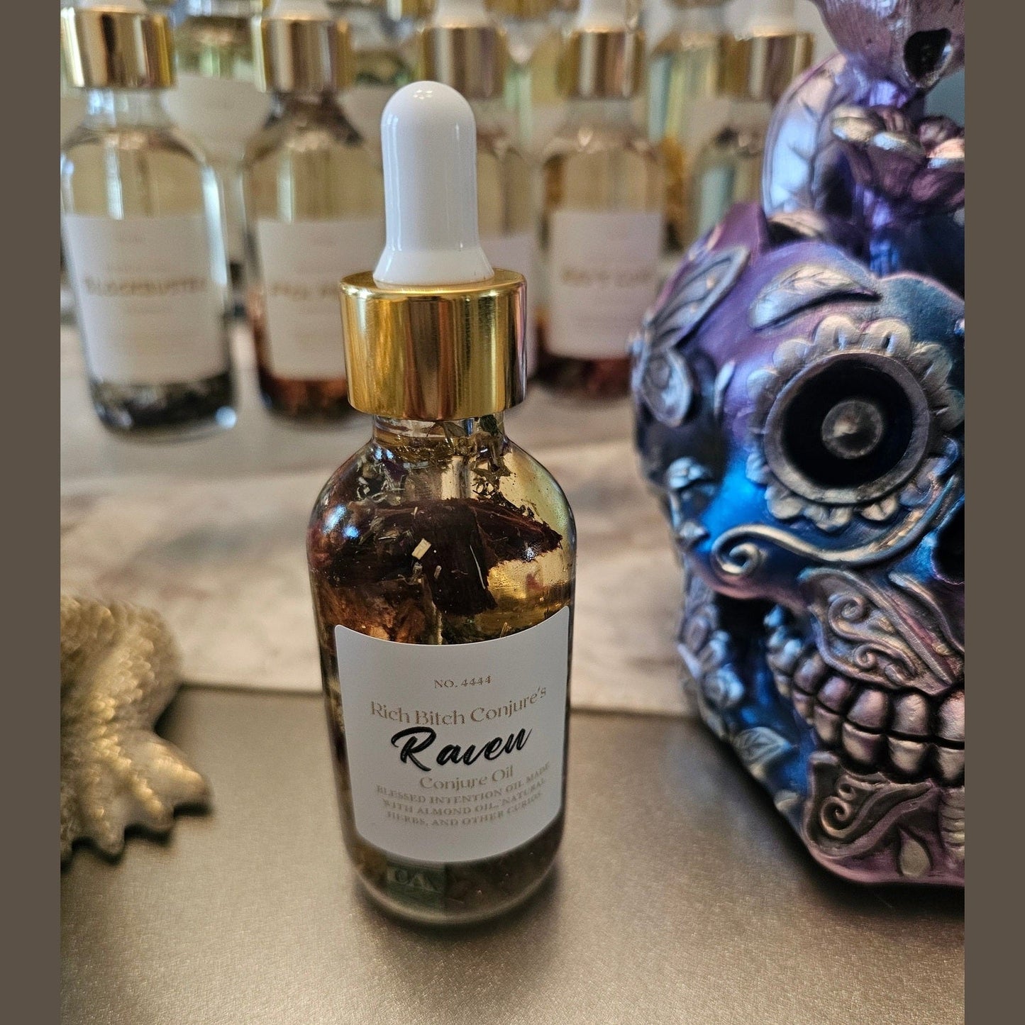 Bottle of 'Raven' cosmetic oil with dropper on a reflective surface, surrounded by decorative items. Raven Hoodoo Conjure Oil for Intuition, Manifestation, & Spiritual Power Ritual Oil