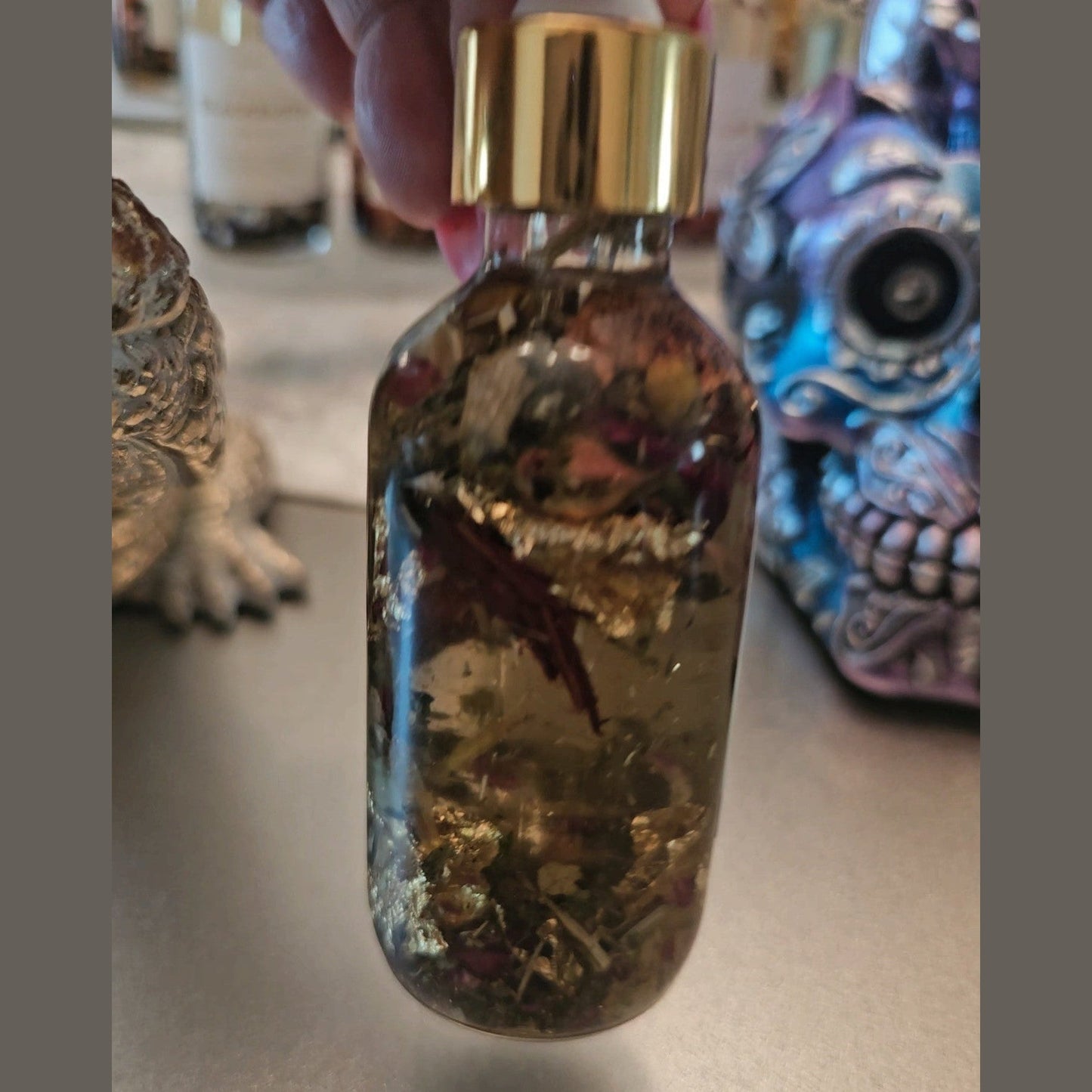 Glass bottle with gold cap held by a hand, with decorative items in the background. Raven Hoodoo Conjure Oil for Intuition, Manifestation, & Spiritual Power Ritual Oil