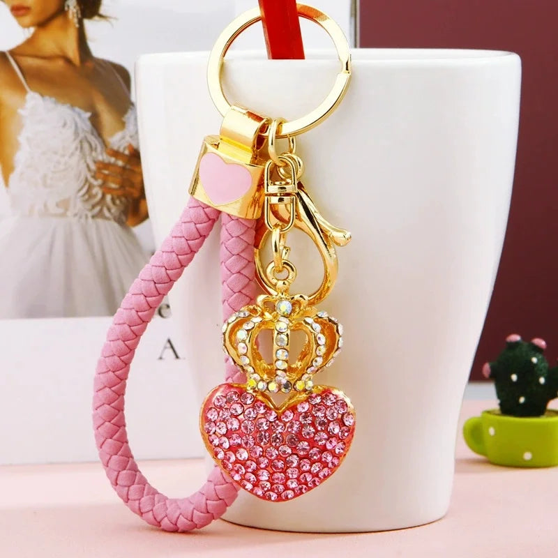 Pink rhinestone heart and crown keychain with a braided pink wrist strap, gold-tone hardware, and lobster clasp displayed hanging from a white tumbler. Stylish women's bag charm and car key holder.