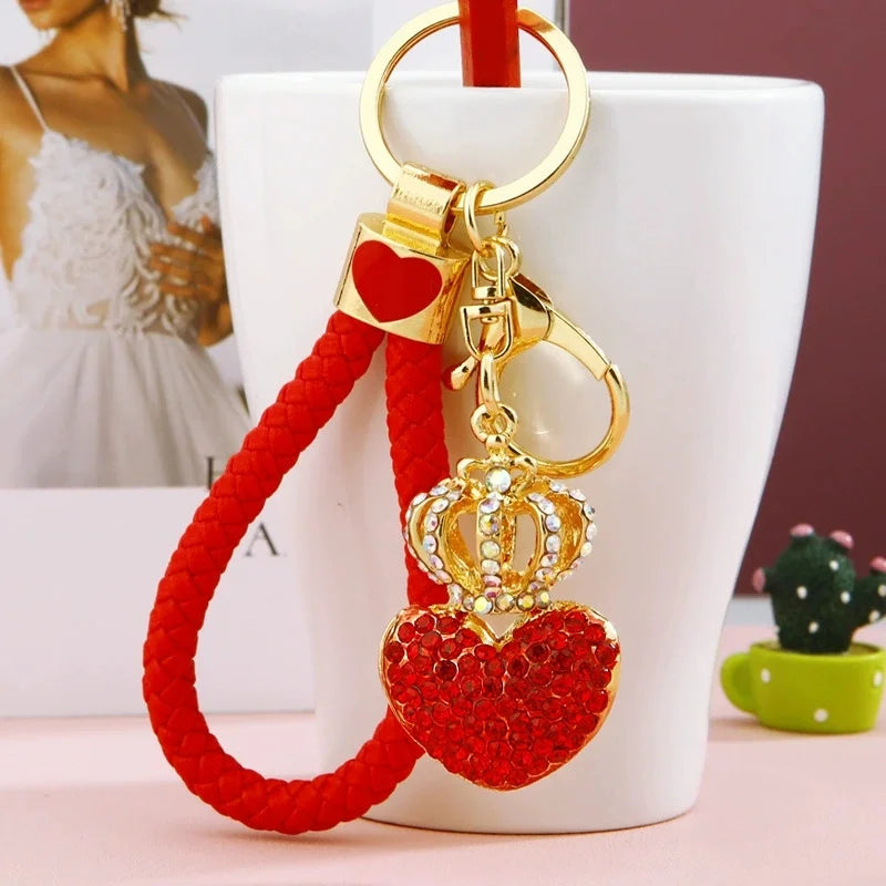 Red rhinestone heart keychain with a gold-tone crown charm and braided red wrist strap, displayed hanging from a white mug against a softly blurred background.