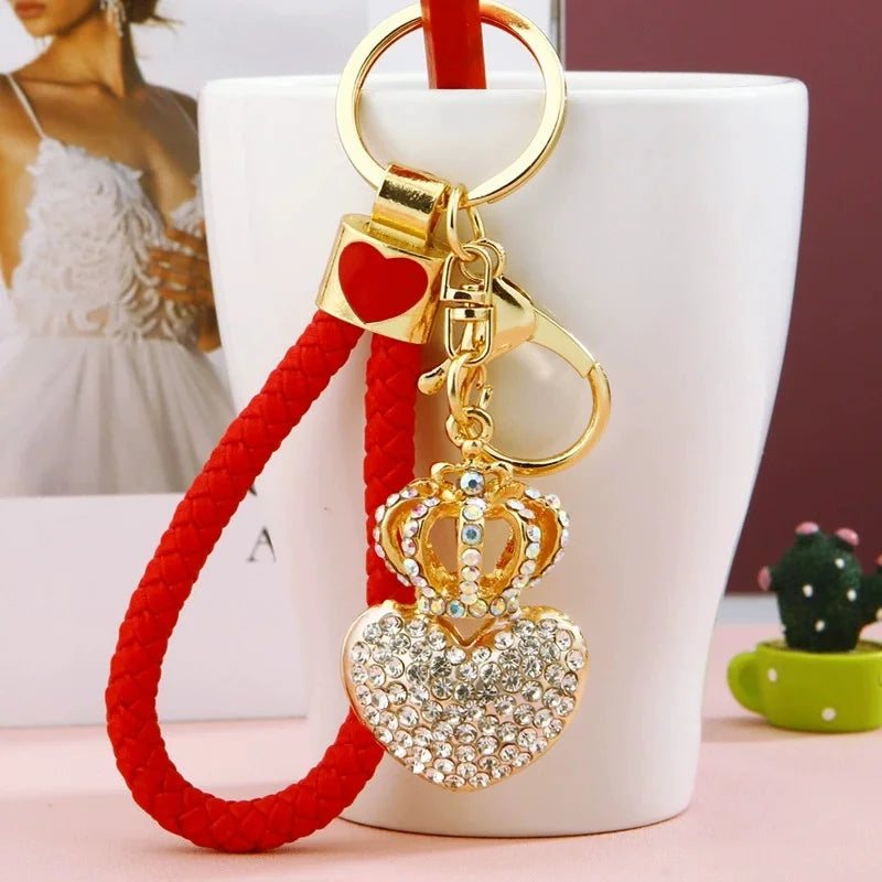 Gold-tone rhinestone crown and heart keychain with a braided red wrist strap, heart-shaped charm, lobster clasp, and key ring, displayed in front of a white mug.