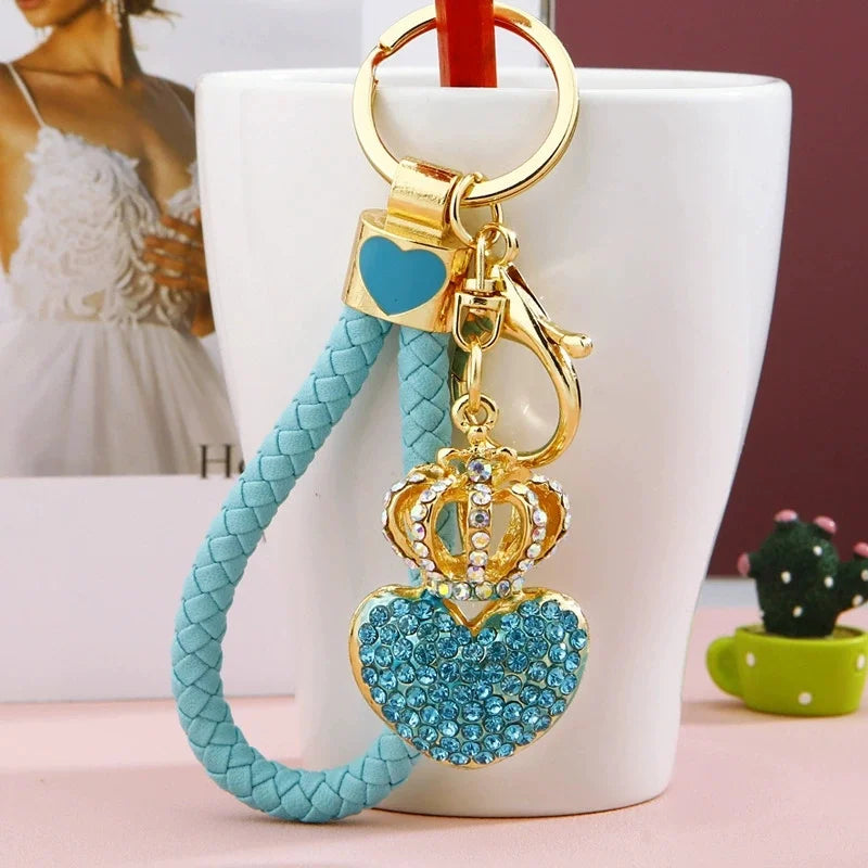 Blue rhinestone heart keychain with a gold-tone crown pendant, braided light blue wrist strap, lobster clasp, and key ring displayed hanging from a white mug.