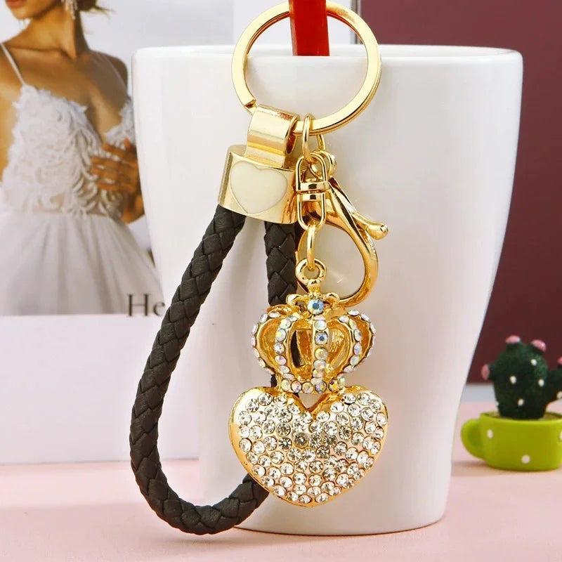 Gold-tone keychain featuring a sparkling rhinestone heart pendant topped with a crown, attached to a black braided wrist strap with a gold key ring and clasp, displayed in front of a white mug on a blurred background.