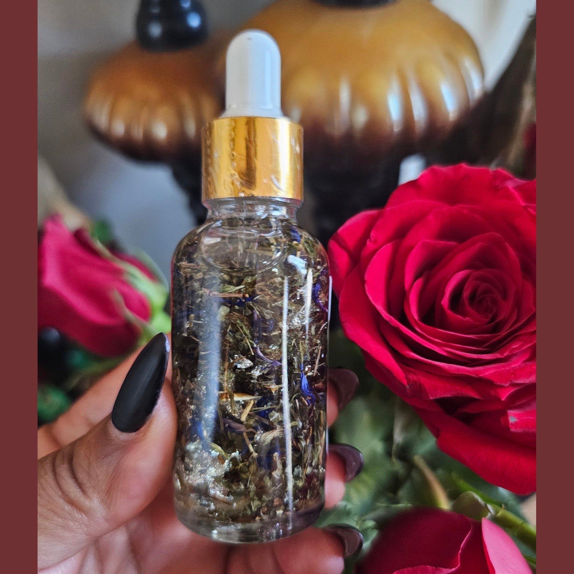 Glass dropper bottle with gold cap held in front of red roses. Rich Bitch Conjure's Blockbuster Open Roads Oil