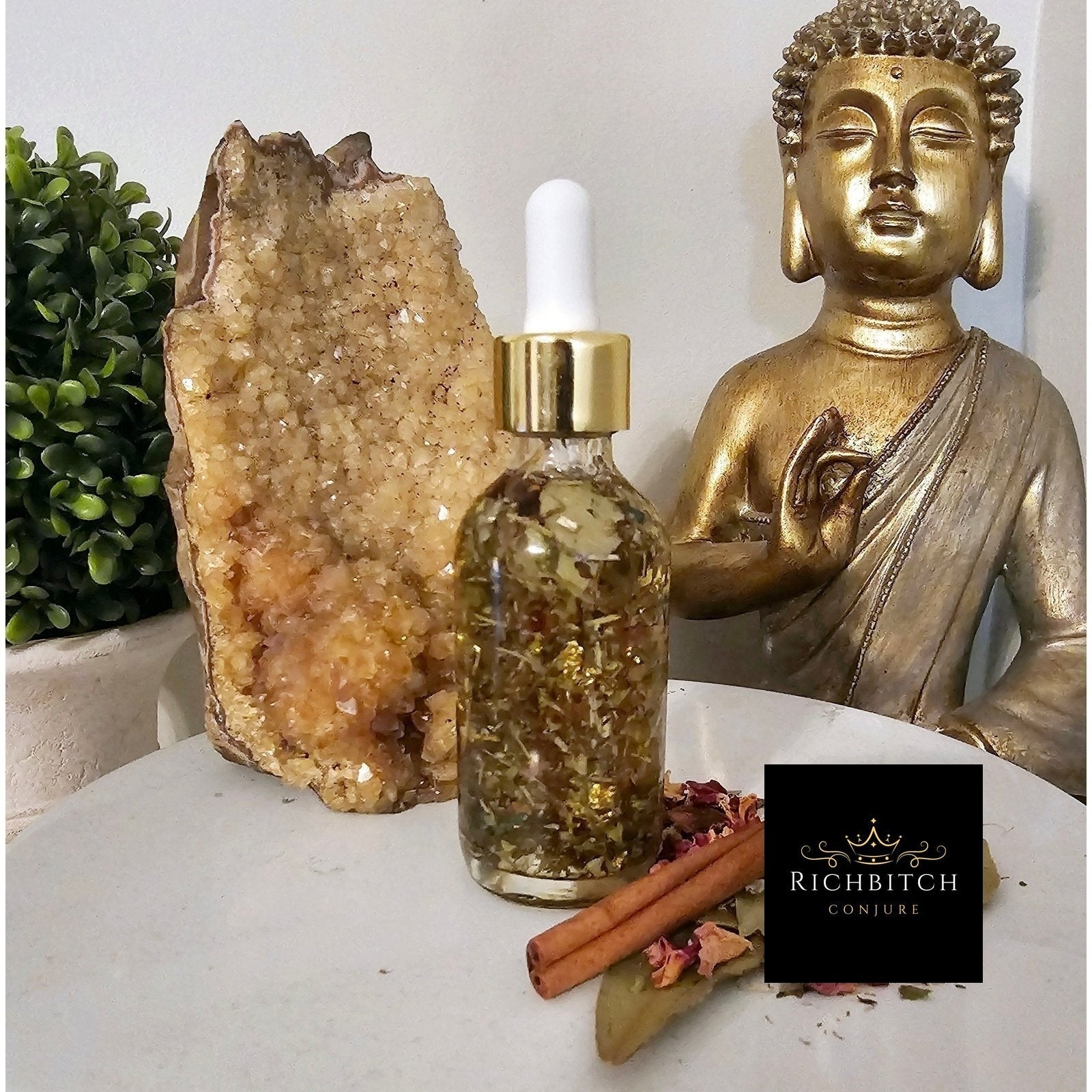 Glass dropper bottle with gold cap on a table with a crystal, cinnamon stick, and Buddha statue. Rich Bitch Conjure's Jasmine Goddess Oil