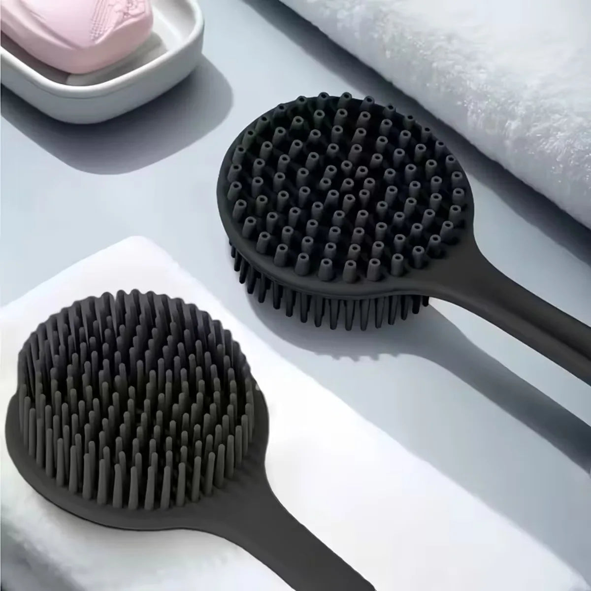 Two black scrubbing brushes on a light gray surface with a white towel and pink soap dish in the background.