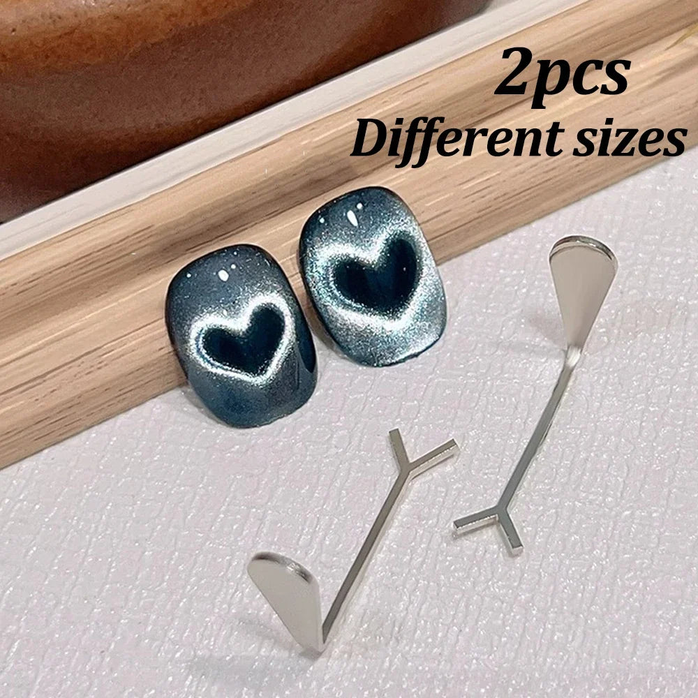 A close-up photograph featuring two pairs of heart-shaped nail art magnets displayed on a combination of rustic wooden texture and white fabric, highlighting the metallic finish and professional design of the tools.