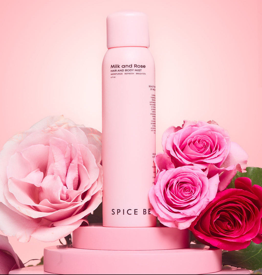 Milk and Rose Hair & Body Mist displayed on a pink pedestal surrounded by blooming pink roses against a soft blush background, showcasing a luxurious floral fragrance and elegant beauty aesthetic.