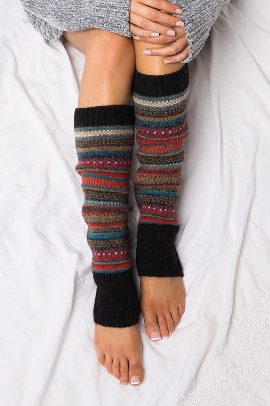 Woman wearing multicolor Fair Isle knit leg warmers featuring a classic Nordic-inspired pattern in red, gray, navy, and cream tones. These soft acrylic leg warmers showcase a thick ribbed cuff and cozy textured knit construction, providing warmth and comfort for cold-weather styling. Perfect for pairing with boots, leggings, jeans, tights, and winter outfits, these fashionable leg warmers add a rustic cabin-chic touch to everyday wear. Ideal for fall and winter fashion, holiday outfits, ski lodge getaways, 