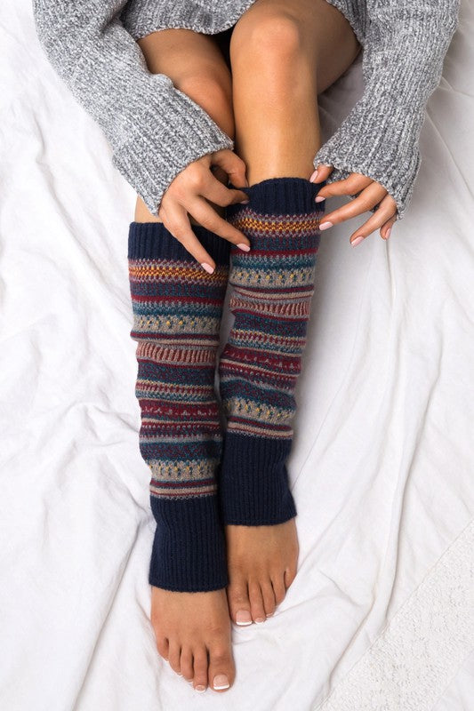 Woman wearing multicolor Fair Isle knit leg warmers featuring a classic Nordic-inspired pattern in red, gray, navy, and cream tones. These soft acrylic leg warmers showcase a thick ribbed cuff and cozy textured knit construction, providing warmth and comfort for cold-weather styling. Perfect for pairing with boots, leggings, jeans, tights, and winter outfits, these fashionable leg warmers add a rustic cabin-chic touch to everyday wear. Ideal for fall and winter fashion, holiday outfits, ski lodge getaways, 