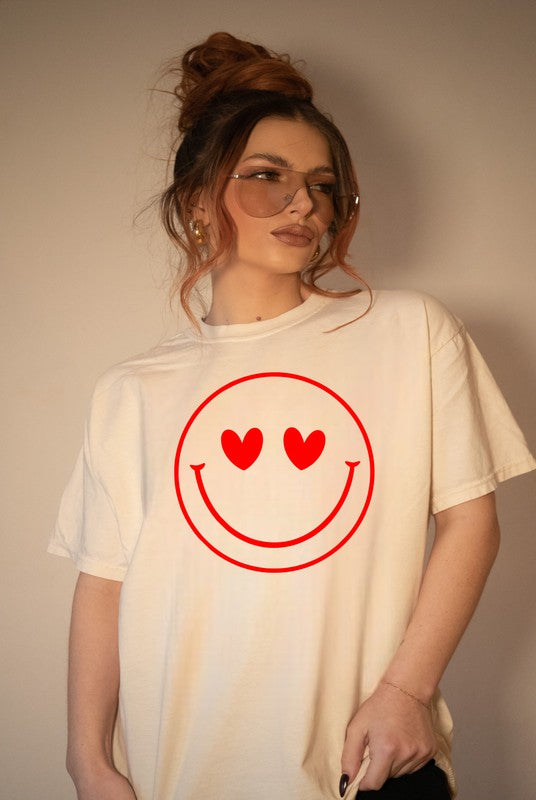 Woman modeling a cream Bella+Canvas unisex graphic T-shirt featuring a large red heart-eyes smiley face design, styled with oversized sunglasses, an effortless messy bun, and a relaxed casual fit against a neutral backdrop.