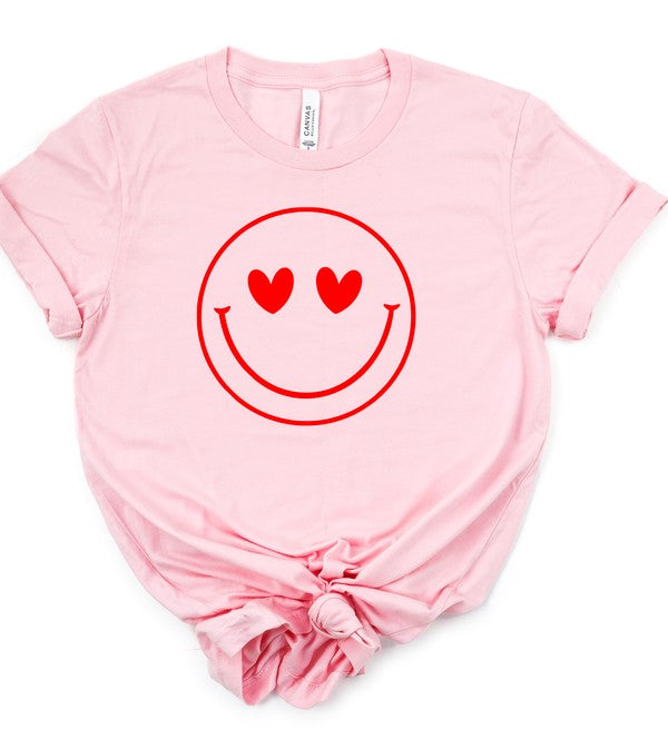 Light pink Bella+Canvas unisex T-shirt featuring a large red smiley face graphic with heart-shaped eyes, displayed with rolled sleeves and a knotted front on a white background.