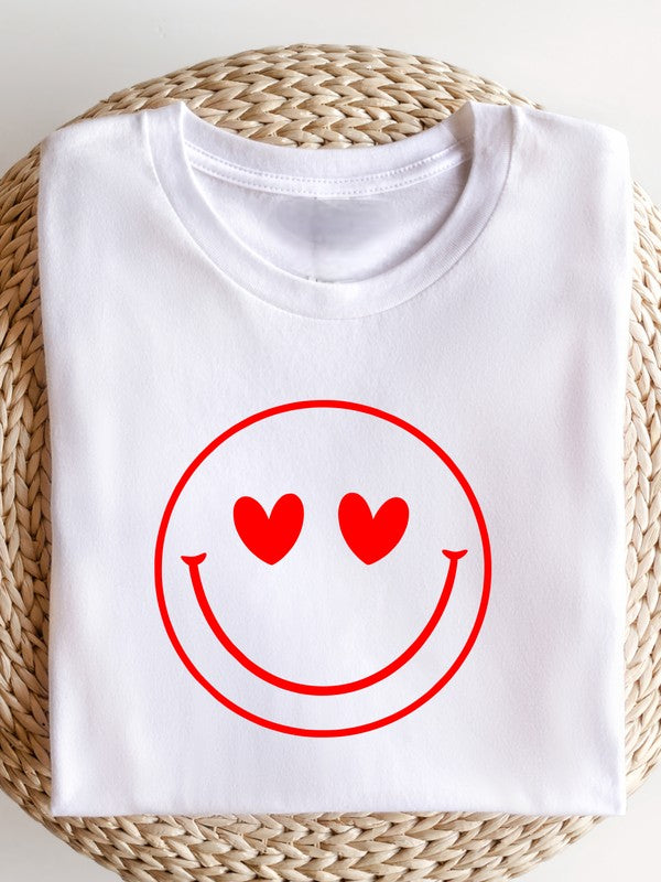 White Bella+Canvas unisex T-shirt folded on a woven straw mat, featuring a bold red smiley face graphic with heart-shaped eyes for a fun, playful, minimalist aesthetic.