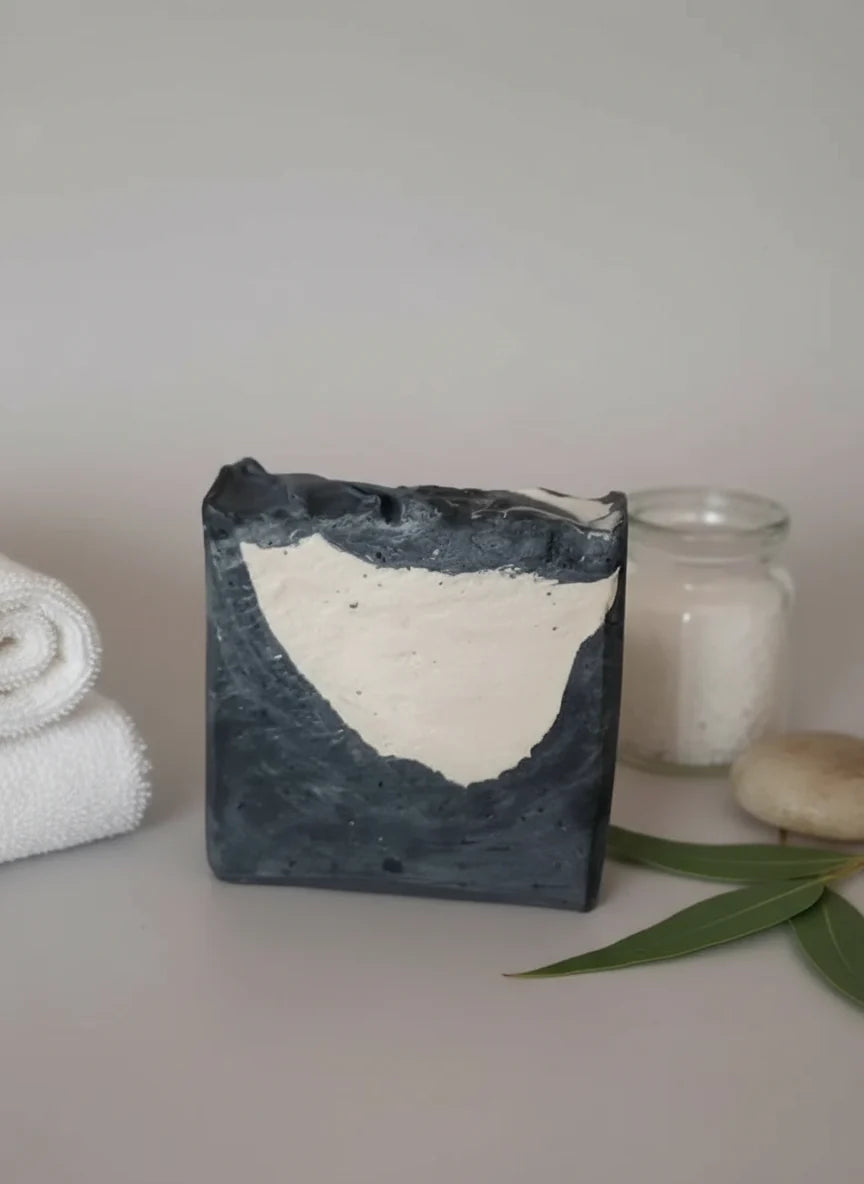Luxurious handmade black-and-white tuxedo-inspired soap bar for men with a masculine design, displayed beside a rolled white towel, candle, and green leaves on a clean spa-style background.
