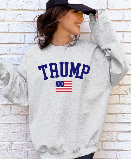 Woman wearing a gray Trump 45/47 USA Flag Graphic Sweatshirt featuring a bold navy "TRUMP" print, American flag graphic, and 45/47 sleeve detail, standing against a white brick wall while wearing a black trucker hat.
