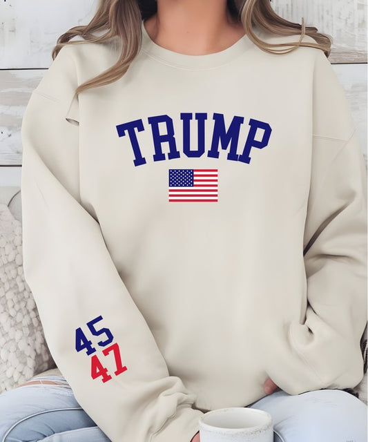Beige sweatshirt with 'TRUMP' and American flag design, worn by a person. Woman wearing a cream beige Trump 45/47 USA Flag Graphic Sweatshirt featuring a bold navy "TRUMP" logo, American flag graphic on the front, and 45/47 print on the left sleeve, posing against a white wood wall.
