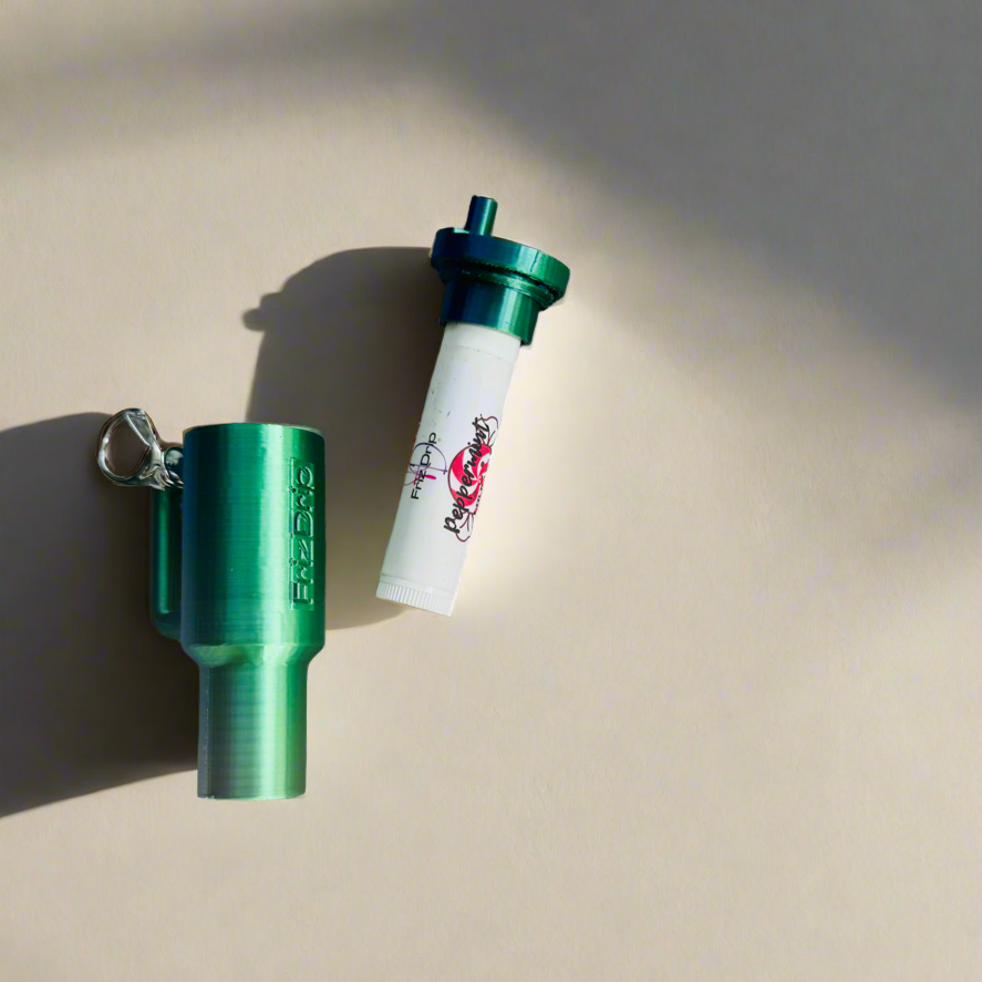 Green tumbler-style silicone lip balm holder keychain with detachable cap and metal key ring, displayed beside a standard lip balm tube on a light background. Reusable purse charm fits most standard lip balm tubes for easy access while traveling or commuting.