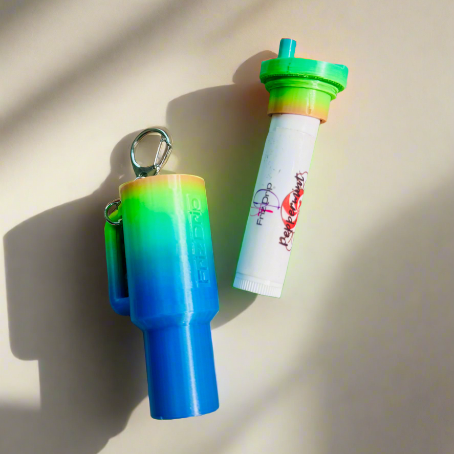 Rainbow gradient tumbler-style silicone lip balm holder keychain displayed beside a standard lip balm tube on a light background.